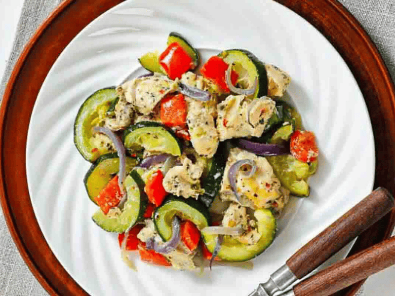 A plate of cooked chicken pieces mixed with zucchini, red bell peppers, and red onion slices, served on a white dish with wooden-handled utensils.
