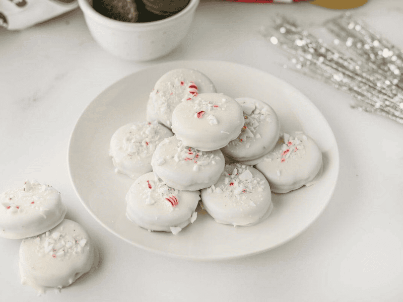 A plate of round cookies coated in white icing, sprinkled with crushed peppermint candies, is displayed on a white surface.
