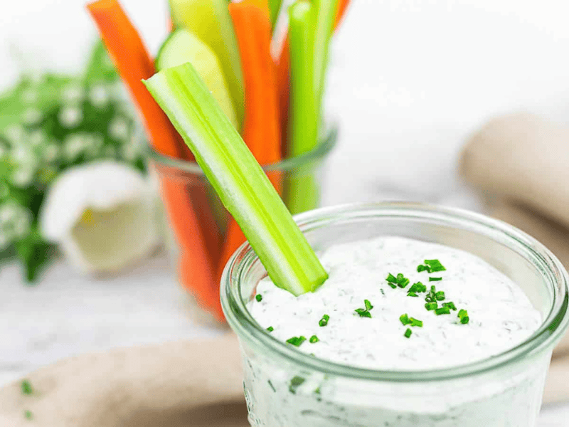 A glass of creamy dip garnished with chopped chives sits in the foreground, with a celery stick inside and a jar of assorted vegetable sticks in the background.