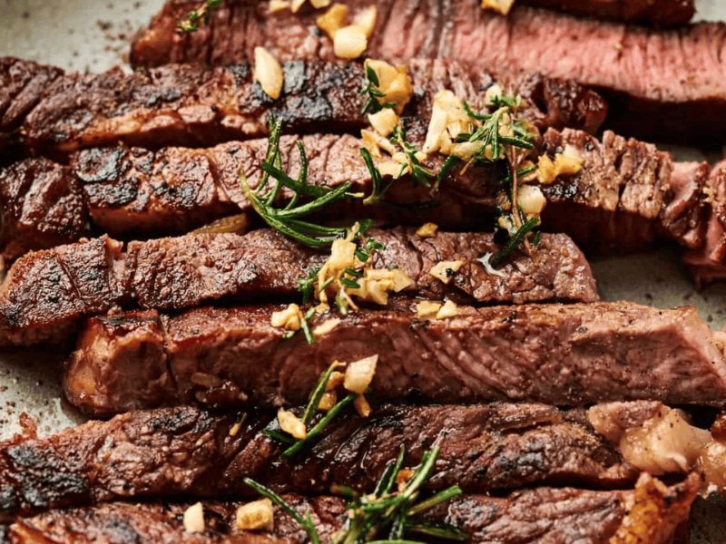 Sliced grilled steak topped with chopped garlic and fresh rosemary on a plate.