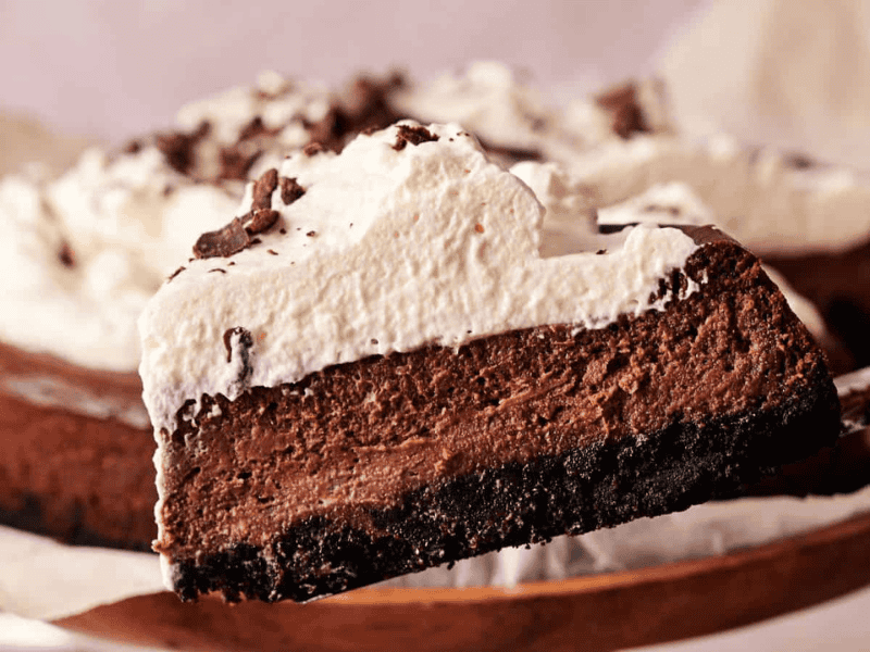 A slice of chocolate cheesecake with a cookie crust and whipped cream topping is being lifted from the whole cake.