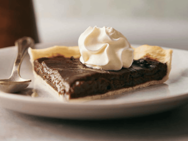 A slice of chocolate pie with a flaky crust, topped with a swirl of whipped cream, sits on a white plate next to a spoon.