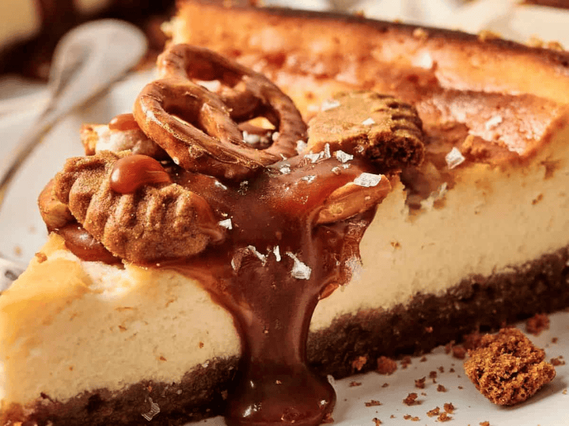 Slice of cheesecake topped with caramel, pretzel, pecan, and sea salt on a plate, with a spoon beside it.