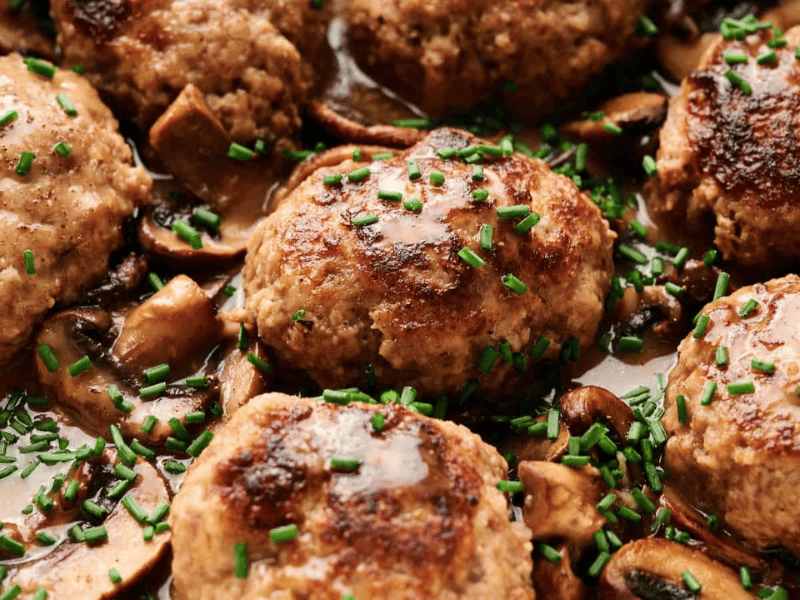 Close-up of browned meat patties with chopped chives and sliced mushrooms in a savory sauce.