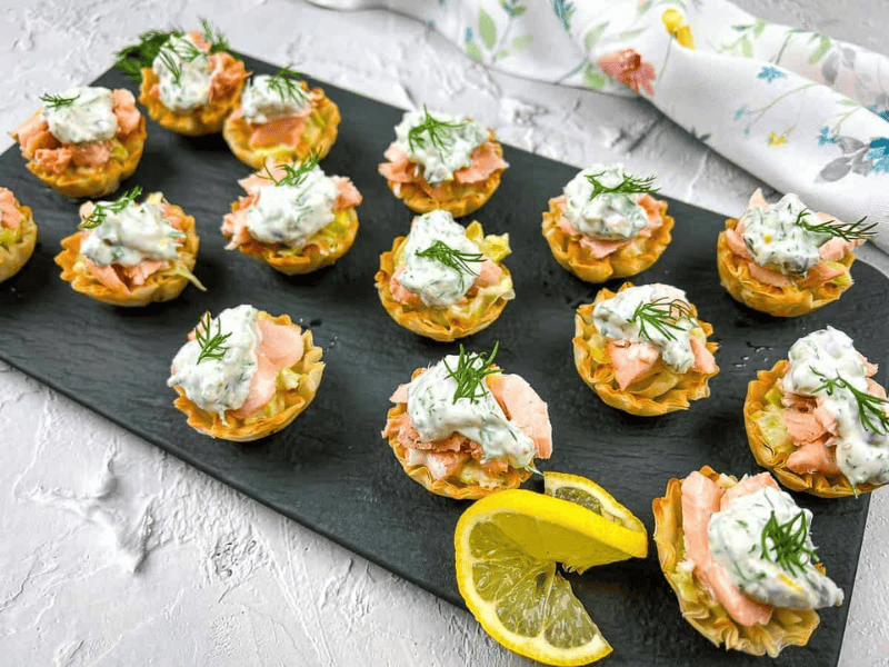 Mini phyllo cups filled with salmon, topped with creamy dill sauce, arranged on a black slate tray with lemon slices and a floral napkin in the background.