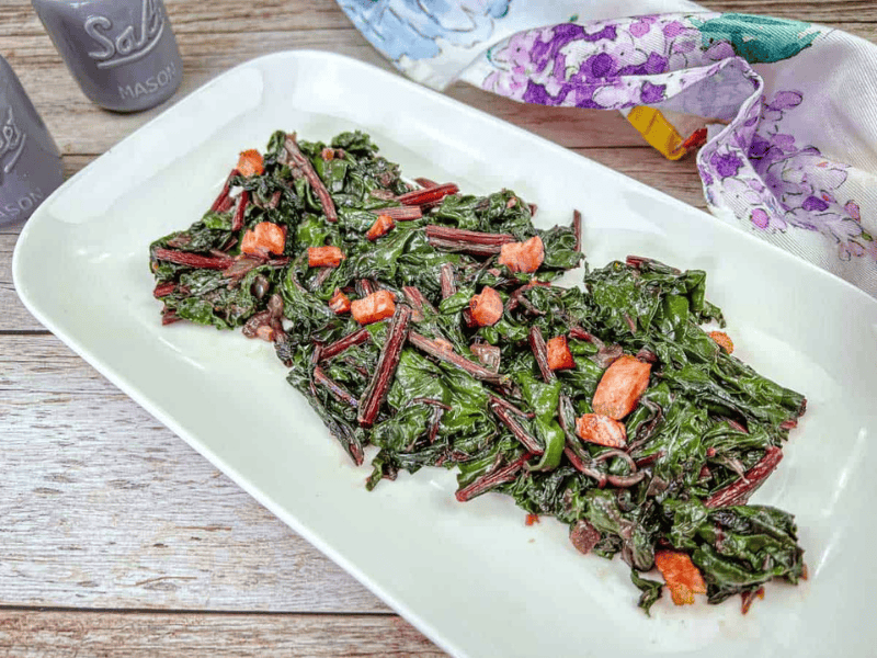Cooked leafy greens with diced red stems and small pieces of bacon are arranged on a white rectangular plate, set on a wooden table with floral cloth nearby.