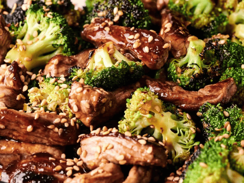 Close-up of stir-fried beef strips and broccoli florets topped with sesame seeds and coated in a glossy sauce.