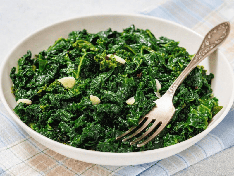 A white bowl filled with saut&eacute;ed kale and sliced garlic, with a fork resting inside, on a checkered cloth.