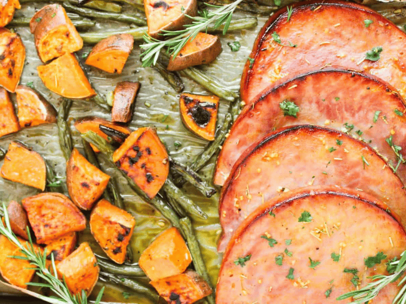 Sliced glazed ham, roasted sweet potato chunks, and green beans arranged on a baking sheet, garnished with rosemary and parsley.
