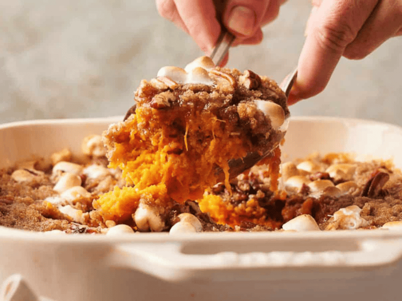 A hand serves a portion of sweet potato casserole topped with marshmallows and pecans from a baking dish.