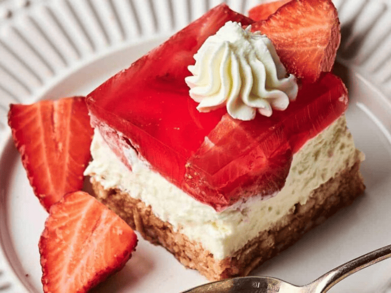A dessert bar with a crumbly base, creamy middle layer, red gelatin topping with strawberries, a dollop of whipped cream, and fresh strawberry slices on a white plate.