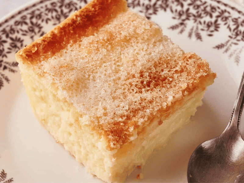 A square slice of sugar-topped custard cake sits on a patterned plate next to a metal spoon.