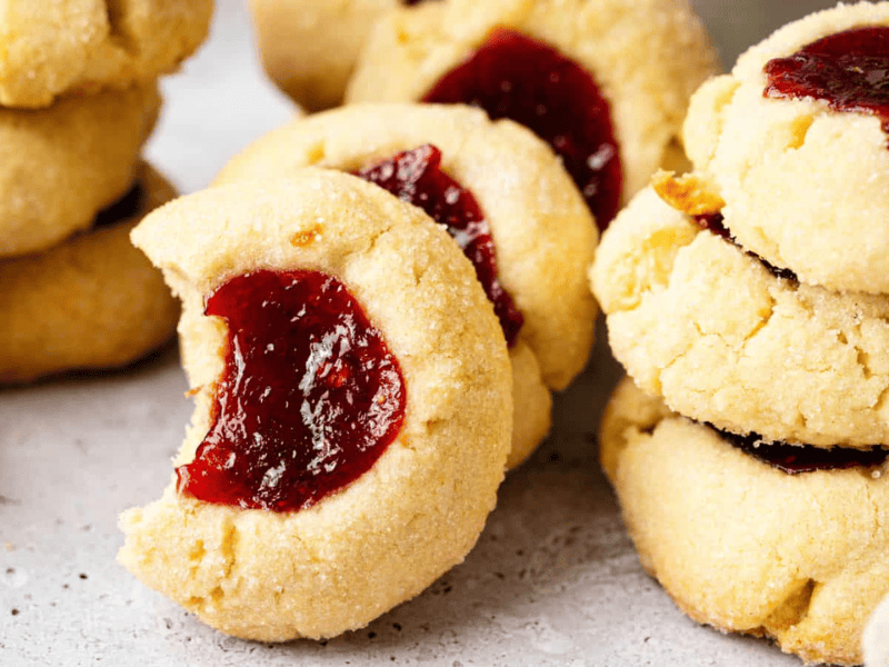 Close-up of several stacks of thumbprint cookies filled with red jam, with one cookie in the foreground showing a bite taken out.