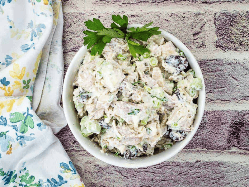 A bowl of chicken salad with chopped celery, green onions, and dark grapes, garnished with parsley, placed on a brick-patterned surface next to a floral napkin.