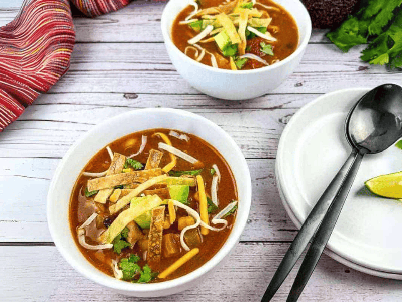 Two bowls of tortilla soup topped with avocado slices, shredded cheese, cilantro, and tortilla strips on a light wooden table with spoons, plates, lime wedges, and a striped cloth nearby.
