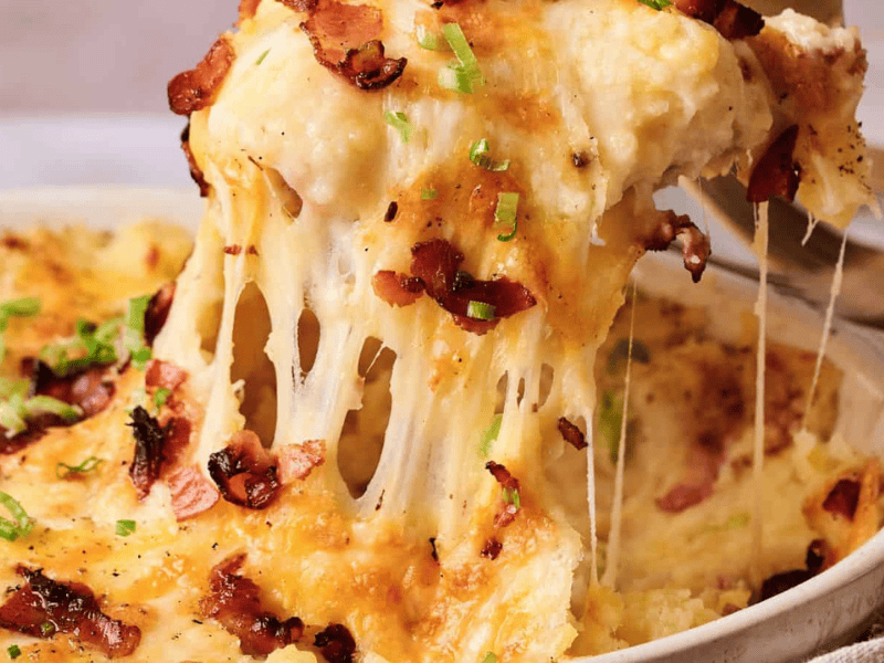 A close-up of a cheesy, baked casserole topped with bacon and chopped green onions, with a serving being lifted out, showing melted cheese stretching.