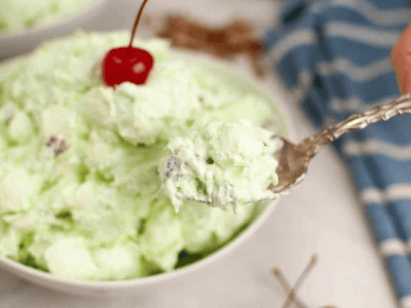 A spoonful of Watergate salad garnished with a cherry, is held over a bowl.