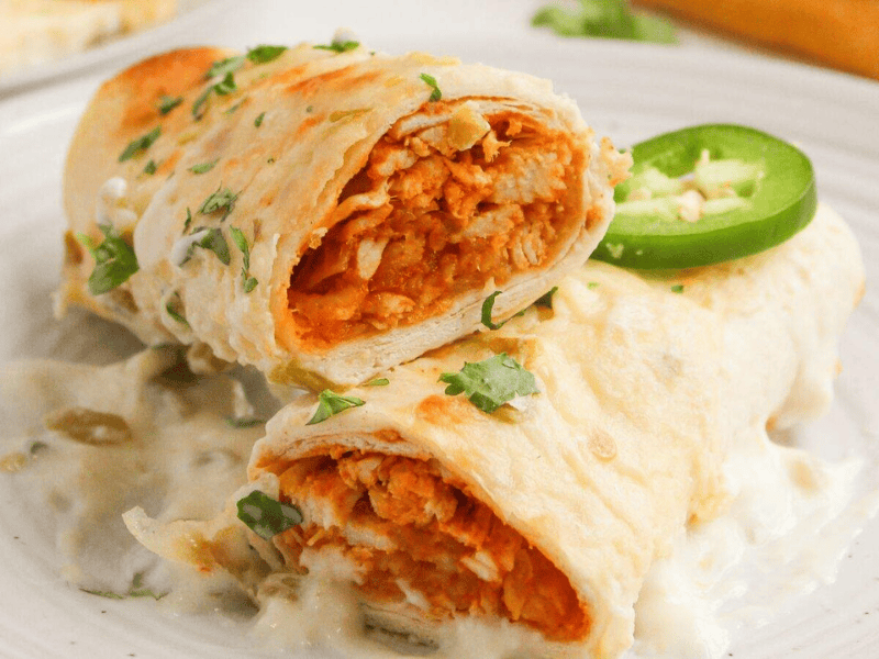 Two tortilla-wrapped enchiladas filled with shredded chicken, topped with creamy white sauce, chopped cilantro, and a jalape&ntilde;o slice, served on a white plate.