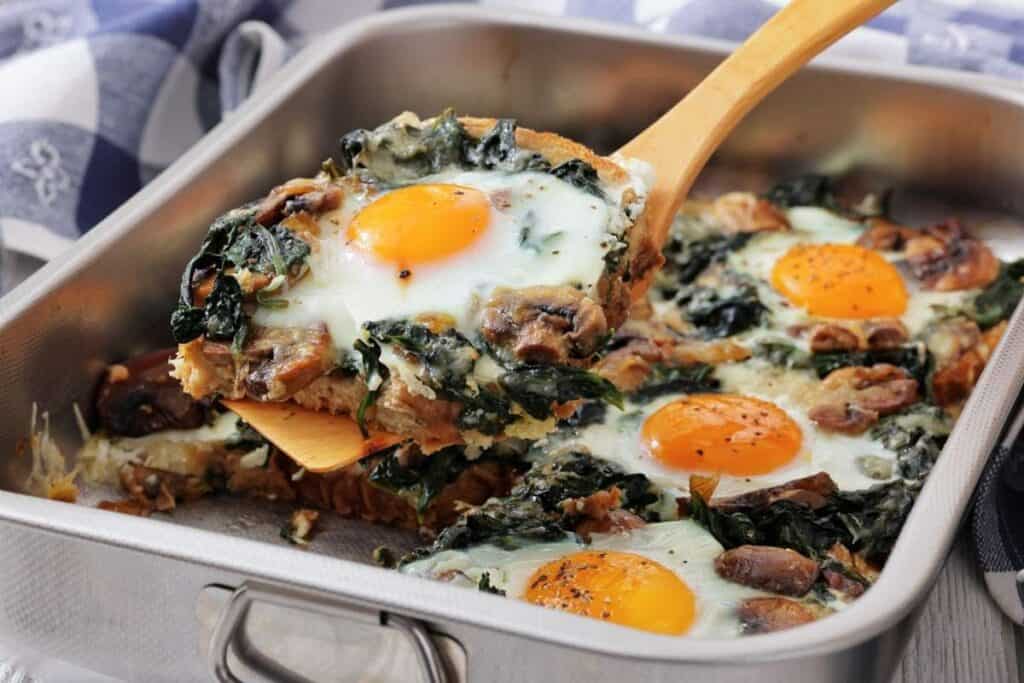 Baked eggs with spinach and mushrooms in a metal baking dish, one portion being lifted with a spatula.