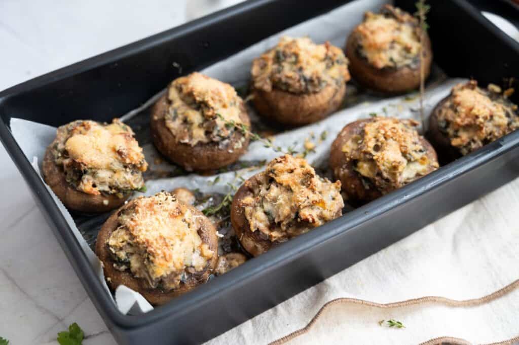 Eight stuffed mushrooms topped with melted cheese are arranged in a black baking dish lined with parchment paper.