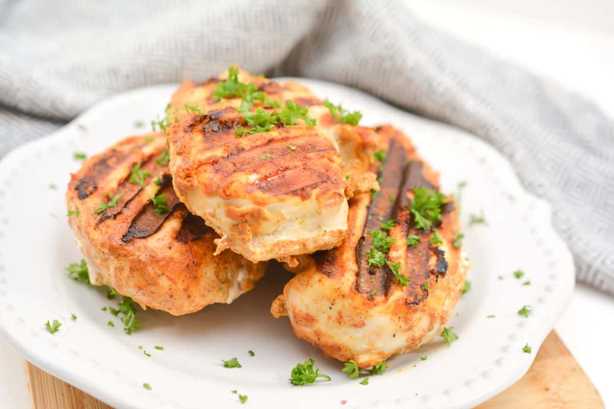 Three grilled chicken breasts garnished with chopped parsley are arranged on a white plate with a gray cloth in the background.