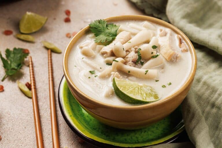 A bowl of creamy soup with noodles, chicken, mushrooms, lime wedge, and cilantro garnish, placed on a green plate with chopsticks and herbs nearby.