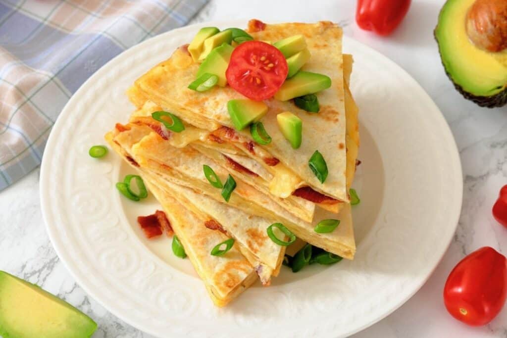A plate of stacked quesadilla wedges topped with diced avocado, sliced green onions, and a cherry tomato slice, with an avocado and tomatoes nearby.
