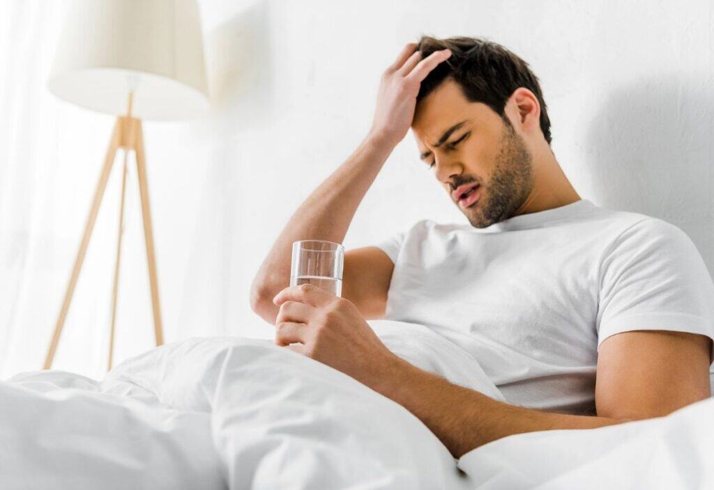 A man sitting up in bed on National Hangover Day holds a glass of water in one hand and touches his forehead with the other, appearing unwell or distressed.