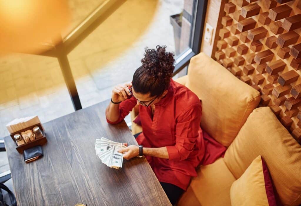 A person in a red shirt sits at a table in a cafe, examining a fan of U.S. dollar bills in their hand.