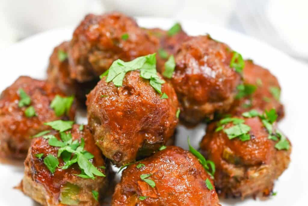 A close-up of several glazed meatballs garnished with chopped fresh parsley on a white plate.