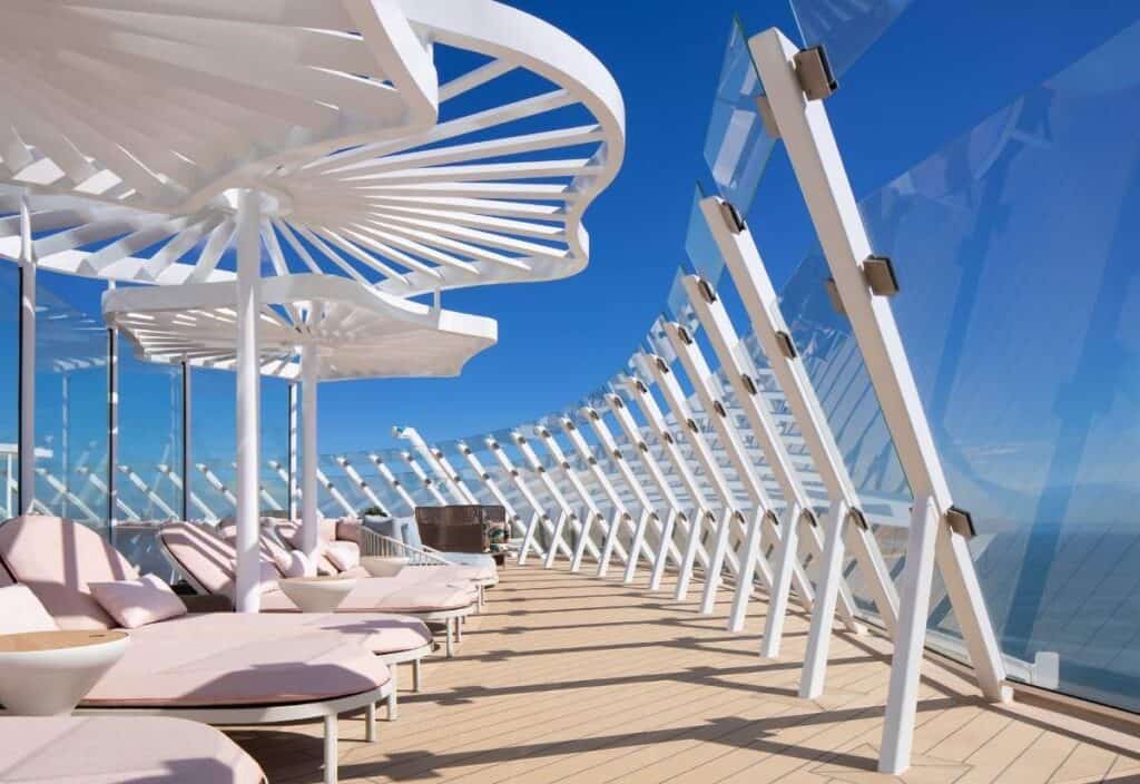Outdoor deck with modern white sunshades, pink lounge chairs, and a glass railing overlooking the ocean under a clear blue sky&mdash;showcasing signature Celebrity Xcel features for an elevated relaxation experience.