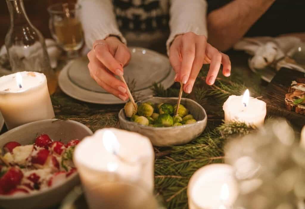 A person uses tongs to serve Brussels sprouts from a bowl on a candlelit dinner table decorated with greenery.
