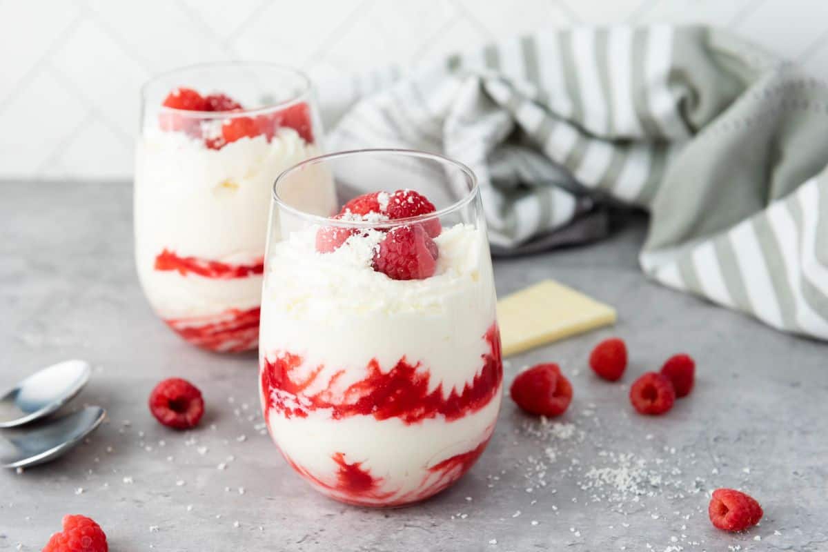 Two glasses filled with layered white cream and red raspberry puree, topped with raspberries and grated chocolate, sit on a gray surface with scattered raspberries and a striped cloth.