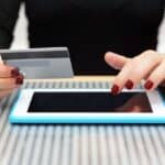 A person holding a credit card in one hand and using the other to tap a tablet on a striped surface, possibly signing up for a subscription model.