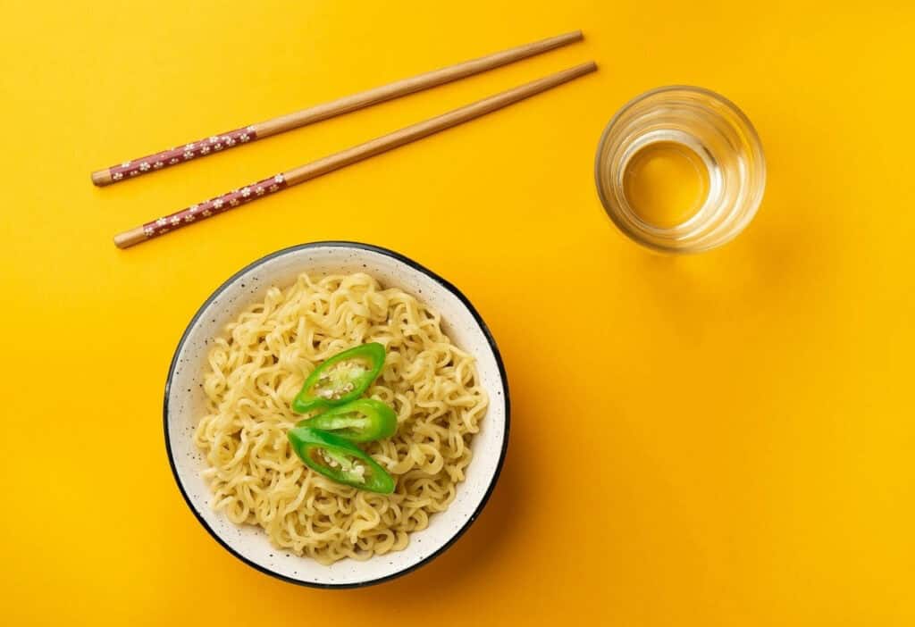 A bowl of cooked instant noodles topped with sliced green chili, a glass of water, and a pair of chopsticks on a yellow background.