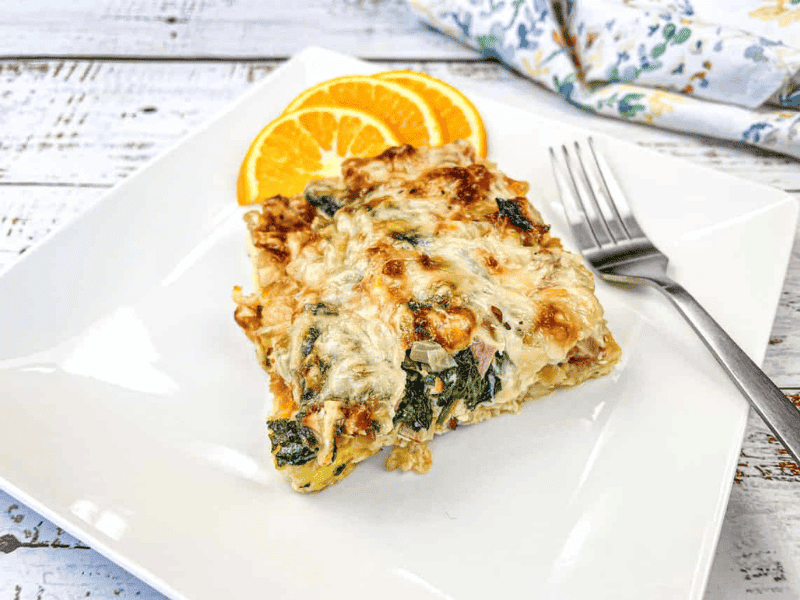 A slice of baked casserole with spinach and melted cheese on a white plate, garnished with orange slices and a fork on the side.