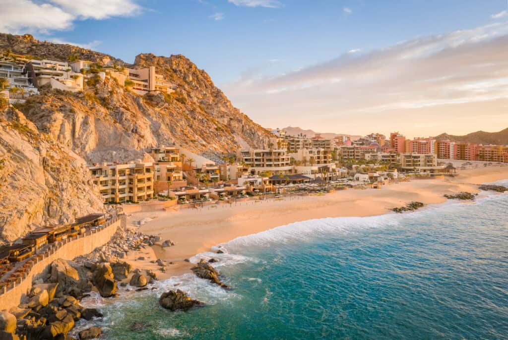 Aerial view of beachfront resorts and buildings along a sandy shore at the base of rocky hills, with gentle ocean waves under a partly cloudy sky at sunset.