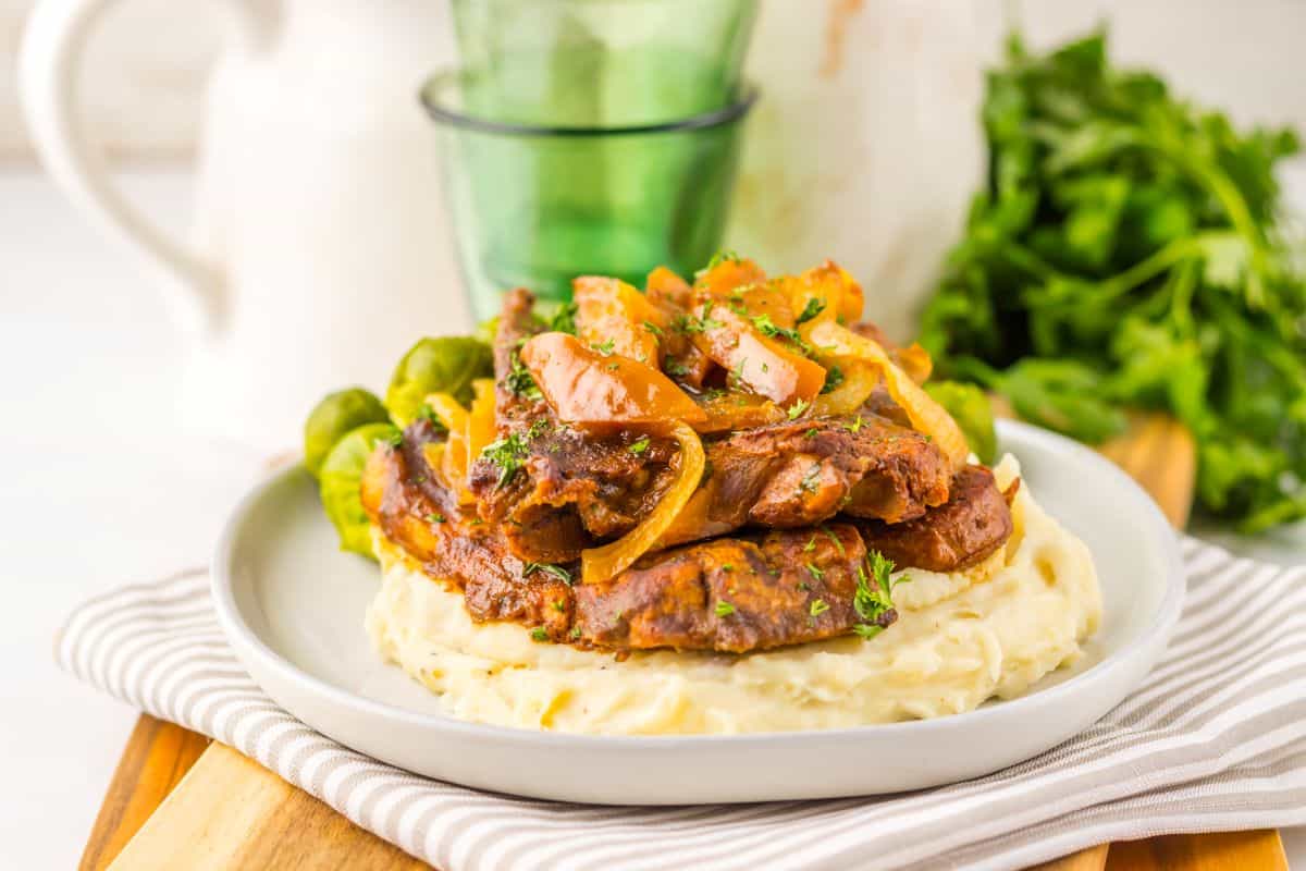 A plate of mashed potatoes topped with slices of cooked meat, saut&eacute;ed onions, and herbs, served with Brussels sprouts.