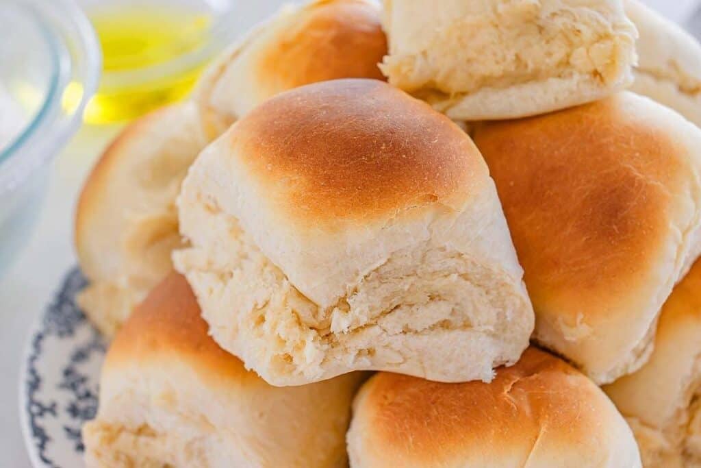 A plate stacked with several golden-brown dinner rolls, these irresistible baked goods showcase a soft, fluffy texture with a slightly shiny top.