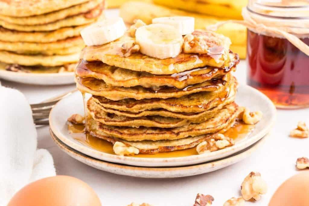 A stack of pancakes topped with banana slices, walnuts, and syrup on a plate offers one of the tastiest breakfast ideas, with eggs and a syrup jar nearby.