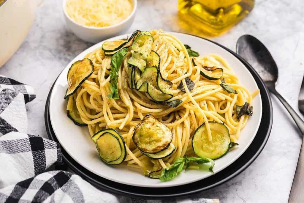 A plate of Pasta Classics: spaghetti topped with sautéed zucchini and fresh basil, with a bowl of grated cheese and a bottle of olive oil in the background.