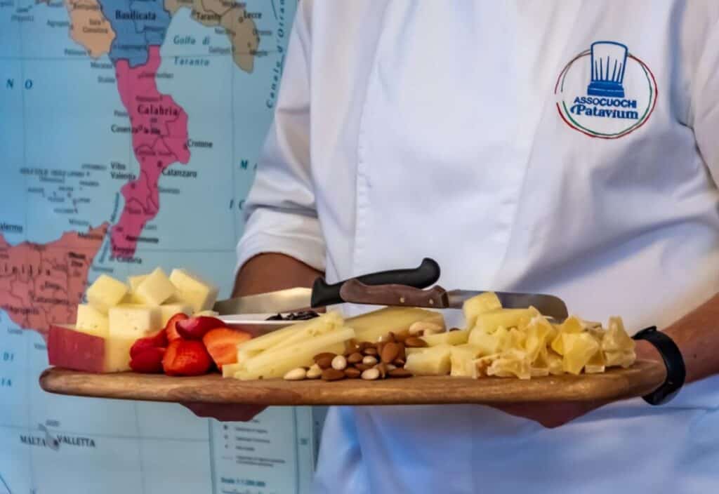 A person in a chef's jacket holds a wooden platter with assorted cheeses, nuts, and strawberries in front of a map of southern Italy, evoking the gourmet experiences found on a luxury barge cruise.