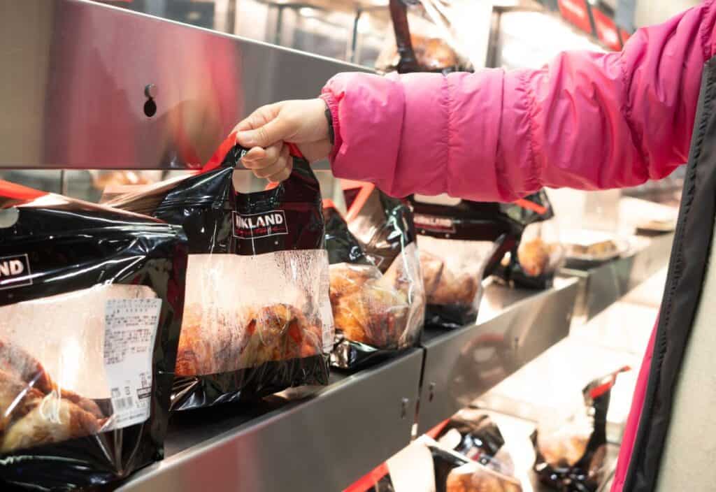 A person wearing a pink jacket selects a packaged rotisserie chicken from a heated display shelf in a grocery store.