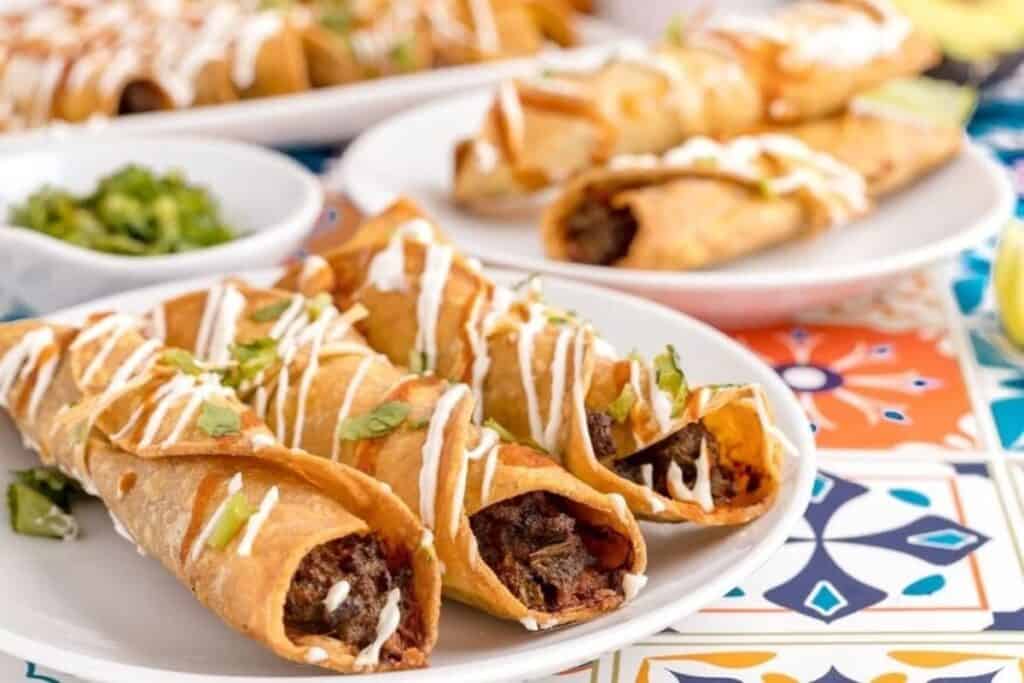 Three beef-filled taquitos topped with sour cream and cilantro are served on a white plate, perfect for easy Weeknight Dinners. More taquitos and a bowl of green salsa can be seen in the background.