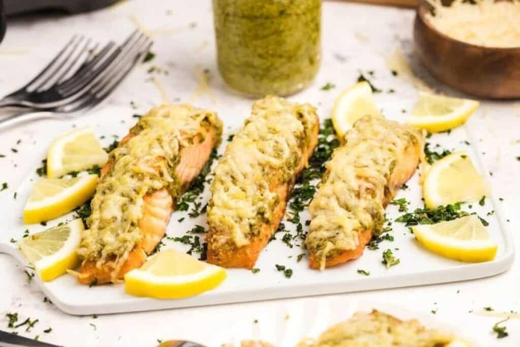 Three baked salmon fillets topped with melted cheese and herbs, garnished with lemon wedges, are arranged on a white rectangular plate&mdash;an ideal choice for effortless weeknight dinners.