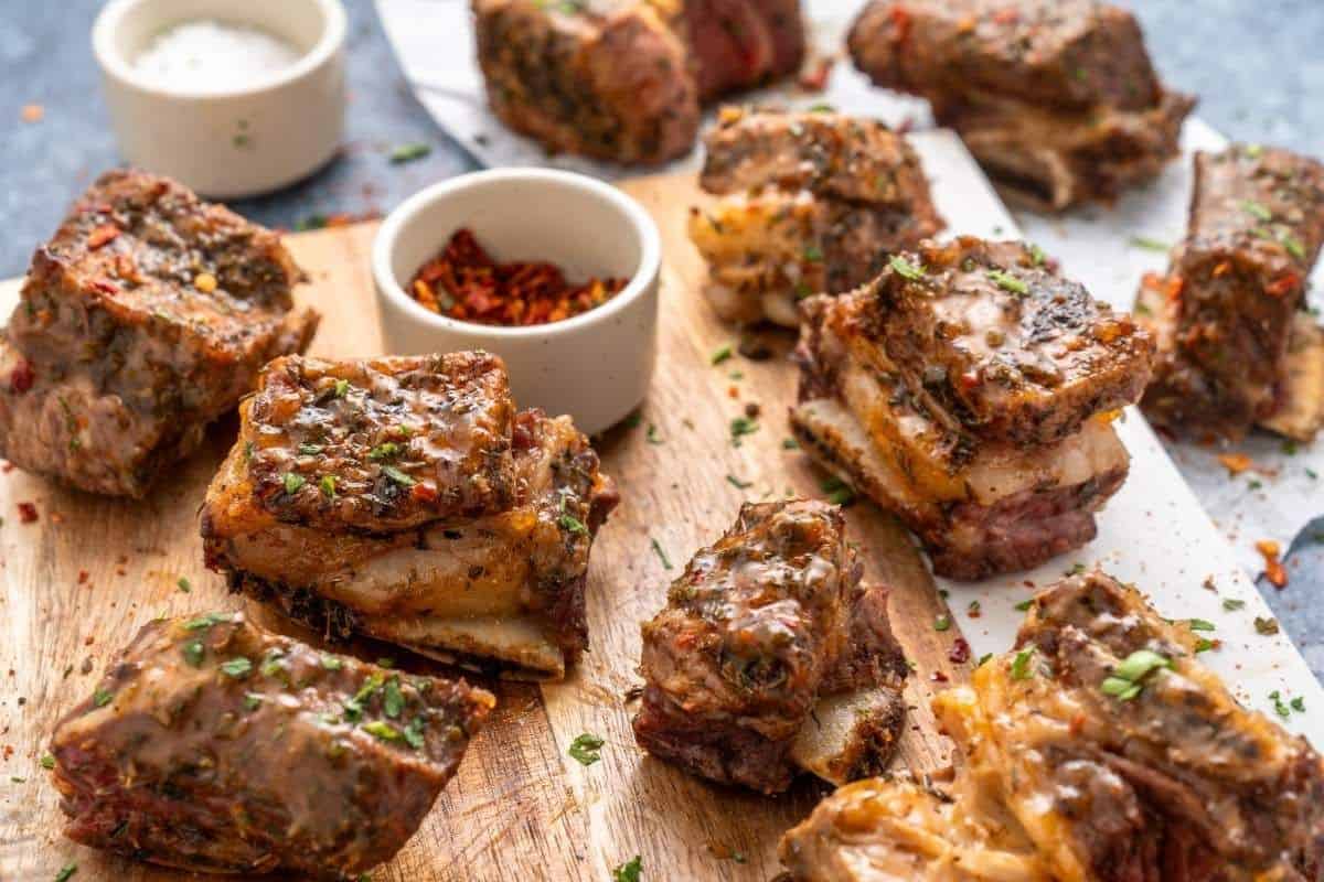 Several pieces of cooked beef short ribs are arranged on a wooden board, garnished with herbs, with small bowls of seasonings in the background—perfect for low effort dinners that don’t sacrifice flavor.