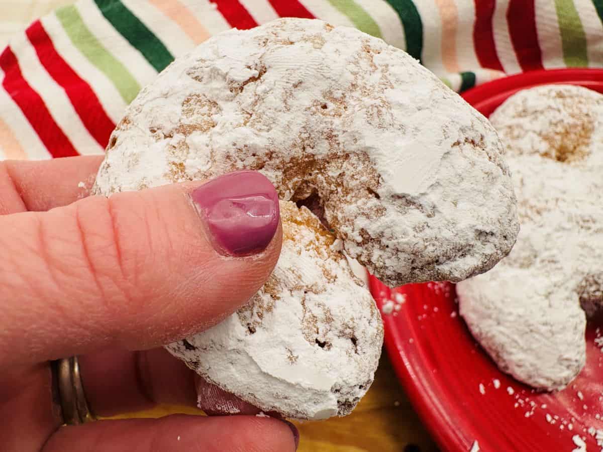 A hand holding a crescent-shaped powdered sugar cookie, with more cookies and a striped cloth visible in the background.