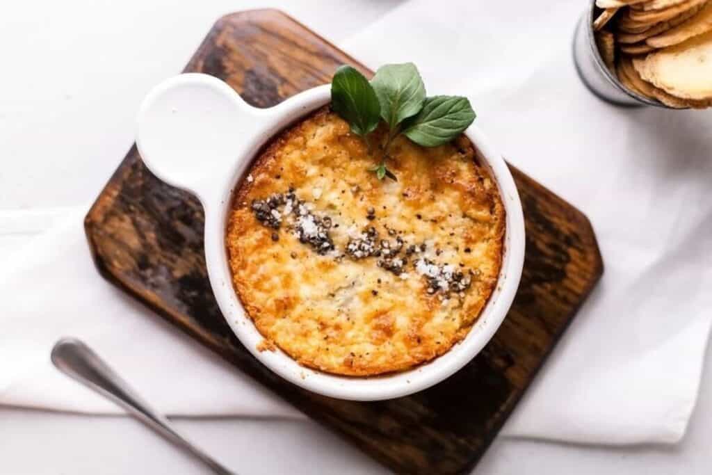 A baked cheese dip garnished with herbs is served in a white ramekin on a wooden board, with a spoon and crackers nearby—perfect for game day appetizers and dips.