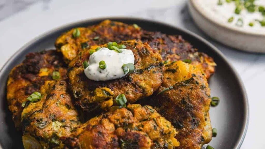 A plate of golden-brown fritters topped with a dollop of creamy sauce and chopped green onions, with a bowl of dip in the background.