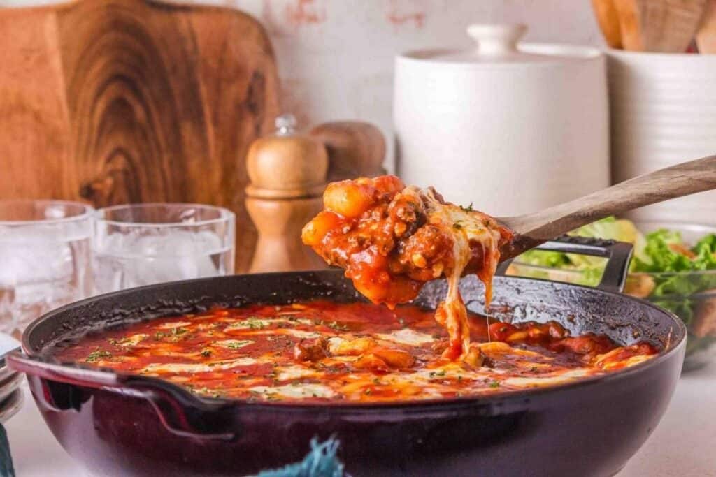 A wooden spoon lifts cheesy baked gnocchi in tomato sauce from a cast iron skillet&mdash;an irresistible addition to your favorite casserole recipes, with kitchen containers and salad in the background.